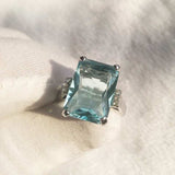 Close-up of a princess aquamarine ring with silver band and side zircon stones on white fabric background